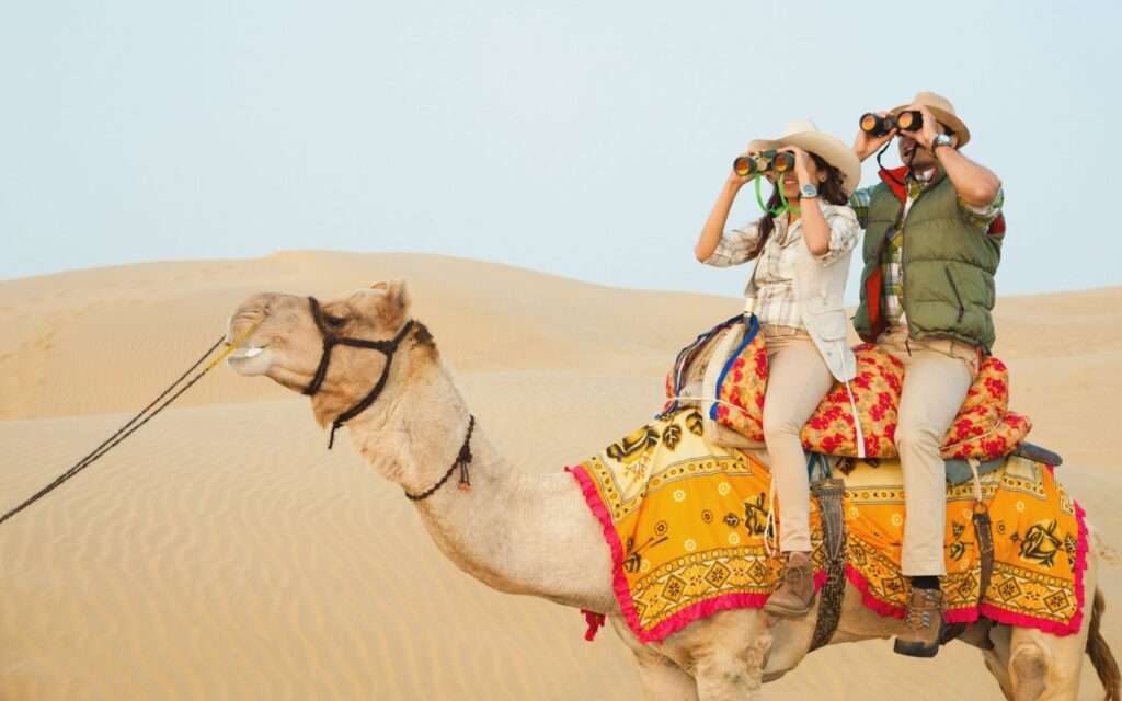 "Two tourists riding on camels in the desert landscape of Pushkar during a camel safari."