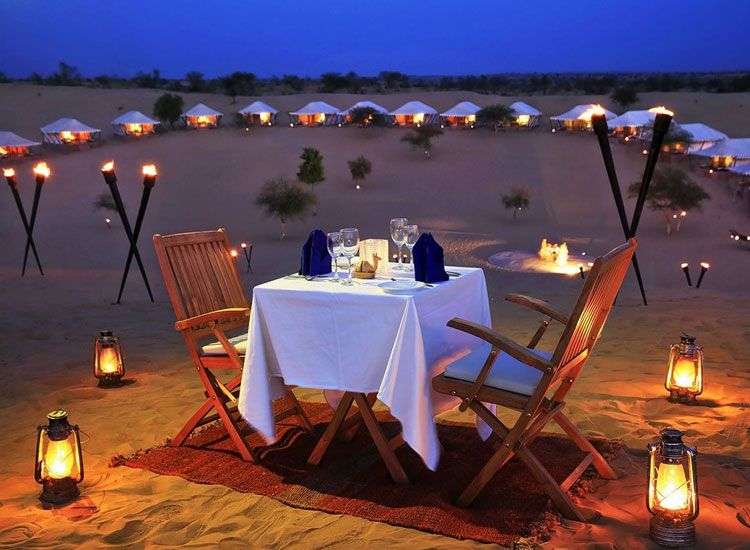 "Two people sitting by a campfire in a desert campsite in Jodhpur, Rajasthan, surrounded by tents and sand dunes in the background."