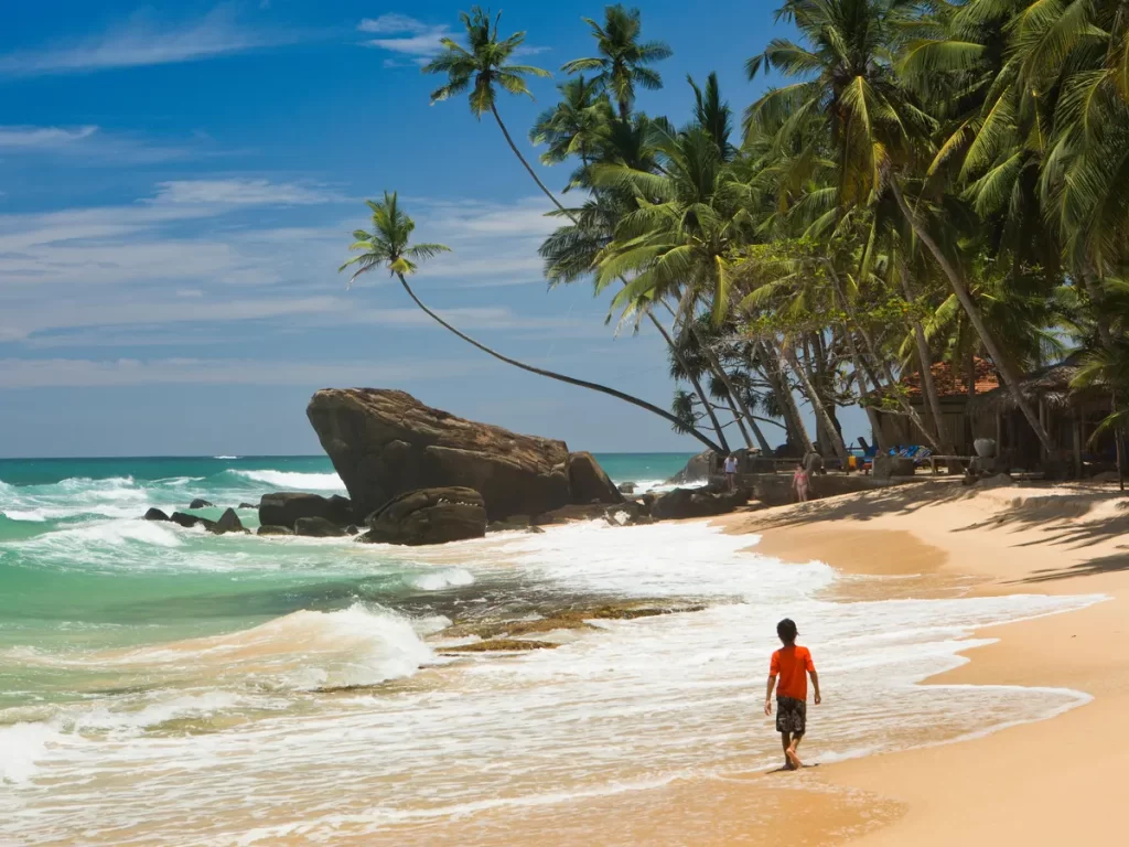 kid walking along the beach of Sri Lanka, with waves splashing around his feet."