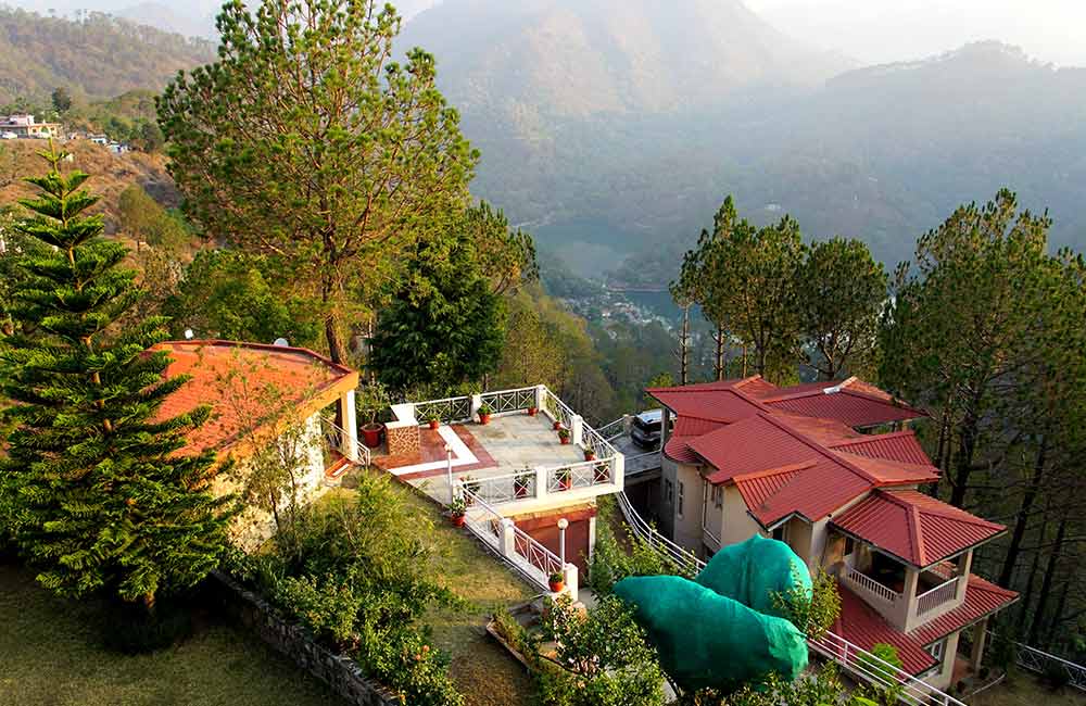 "A view of the serene Bhimtal resort surrounded by lush green trees and a lake in the foreground."