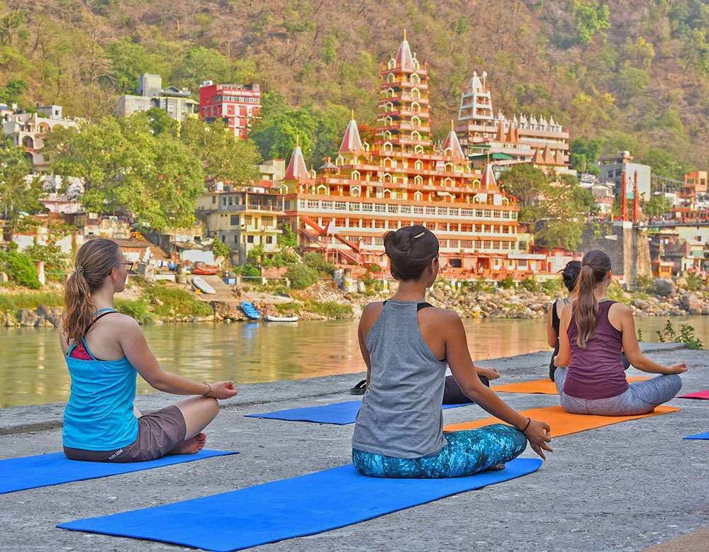 "Girls meditating next to the Rishikesh river with the temple of Rishikesh in the background"