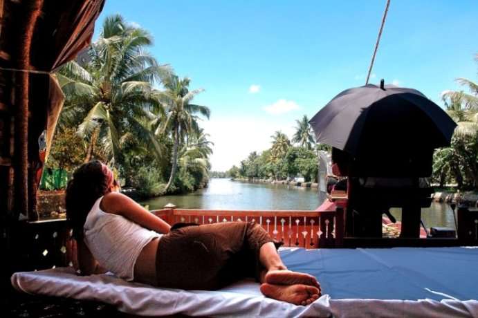 "Relaxing on a houseboat in Kerala's backwaters - a woman lying down on a comfortable couch, enjoying the scenic views of lush greenery and calm waters."