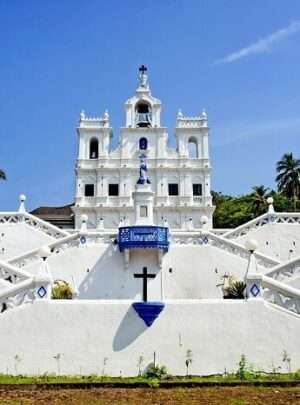 "Exterior view of Mary Immaculate Church in Panjim, Goa, with its tall white spires and ornate facade, against a blue sky with palm trees in the foreground."