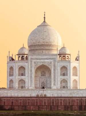 "White marble Taj Mahal mausoleum in Agra, India with a clear blue sky and green trees in the foreground."