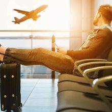 A man sitting in an airport terminal, looking out the window and watching a plane take off.